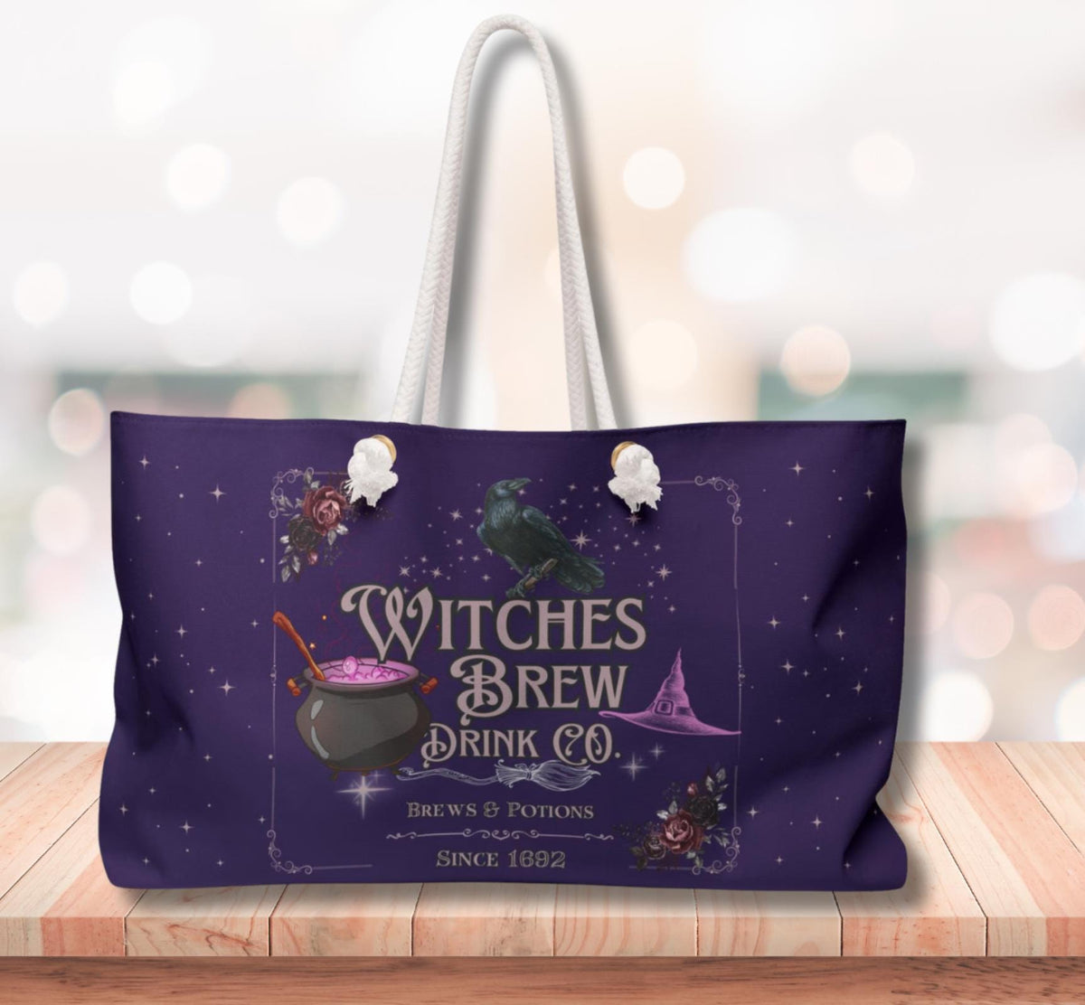 A purple tote bag with white handles, featuring a Halloween-themed graphic that reads 'Witches Brew Drink Co.' with a cauldron, potion, crow, stars, and a blurred festive background.