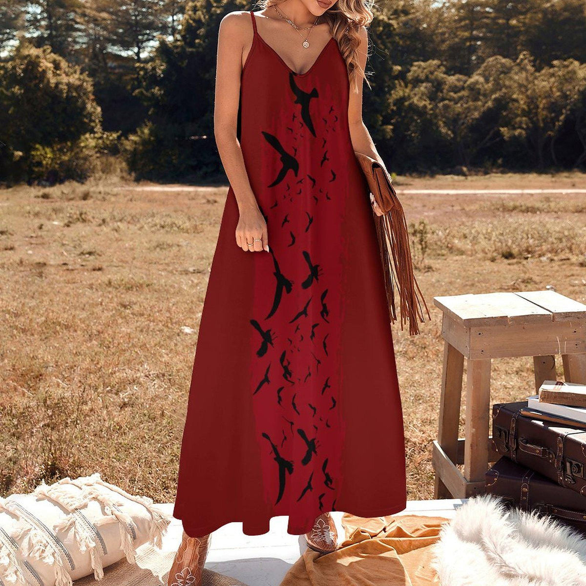A woman wearing a long red maxi dress with black bird silhouettes printed down the front, standing outdoors in a dry grassy field near a wooden bench and suitcase.