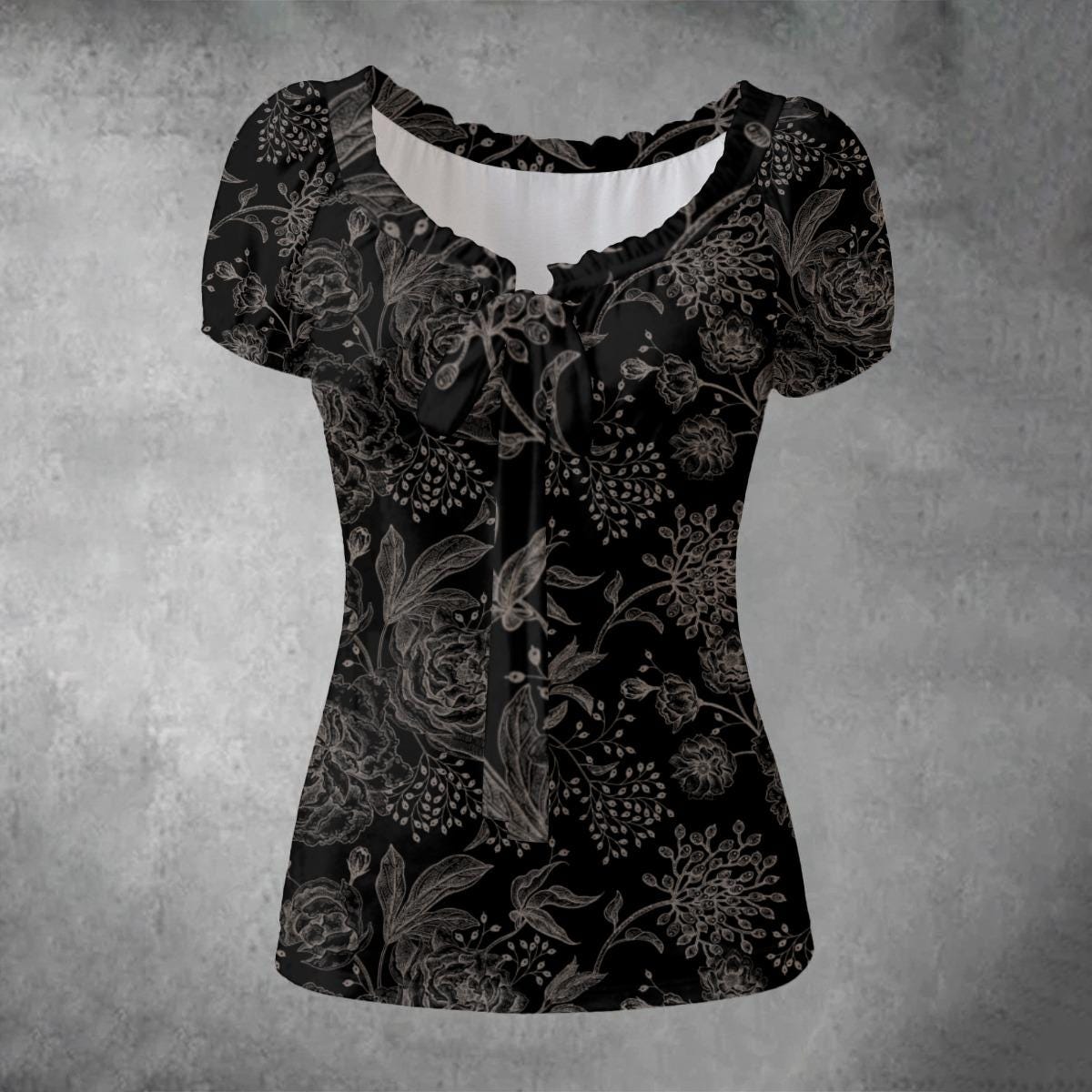 A short-sleeve black blouse with a gray floral and bird pattern, featuring a ruffled scoop neckline and a tie detail at the center chest, displayed against a textured gray background.
