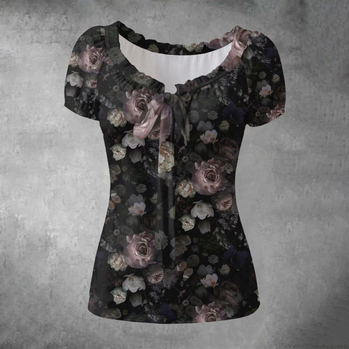 A short-sleeve women's blouse with a ruffled scoop neckline and a front tie detail, featuring an all-over dark floral print with roses in muted pink, gray, and white on a black background, shown against a textured gray backdrop.