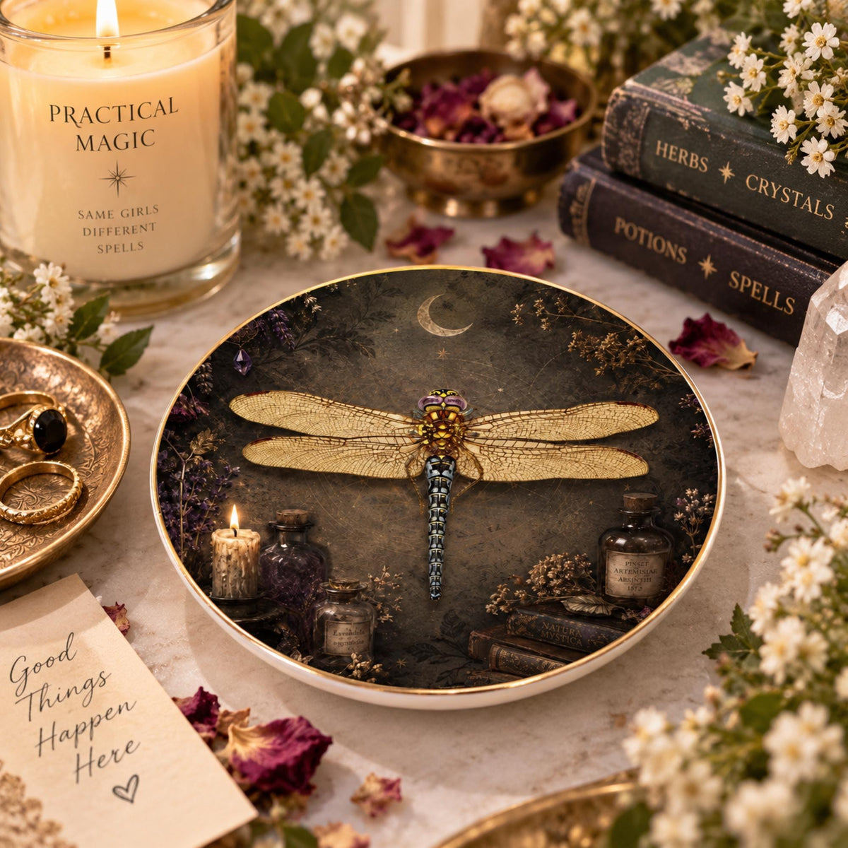 Angled view of a round gold-rimmed dragonfly jewelry dish with a vintage botanical scene beside books and a candle.
